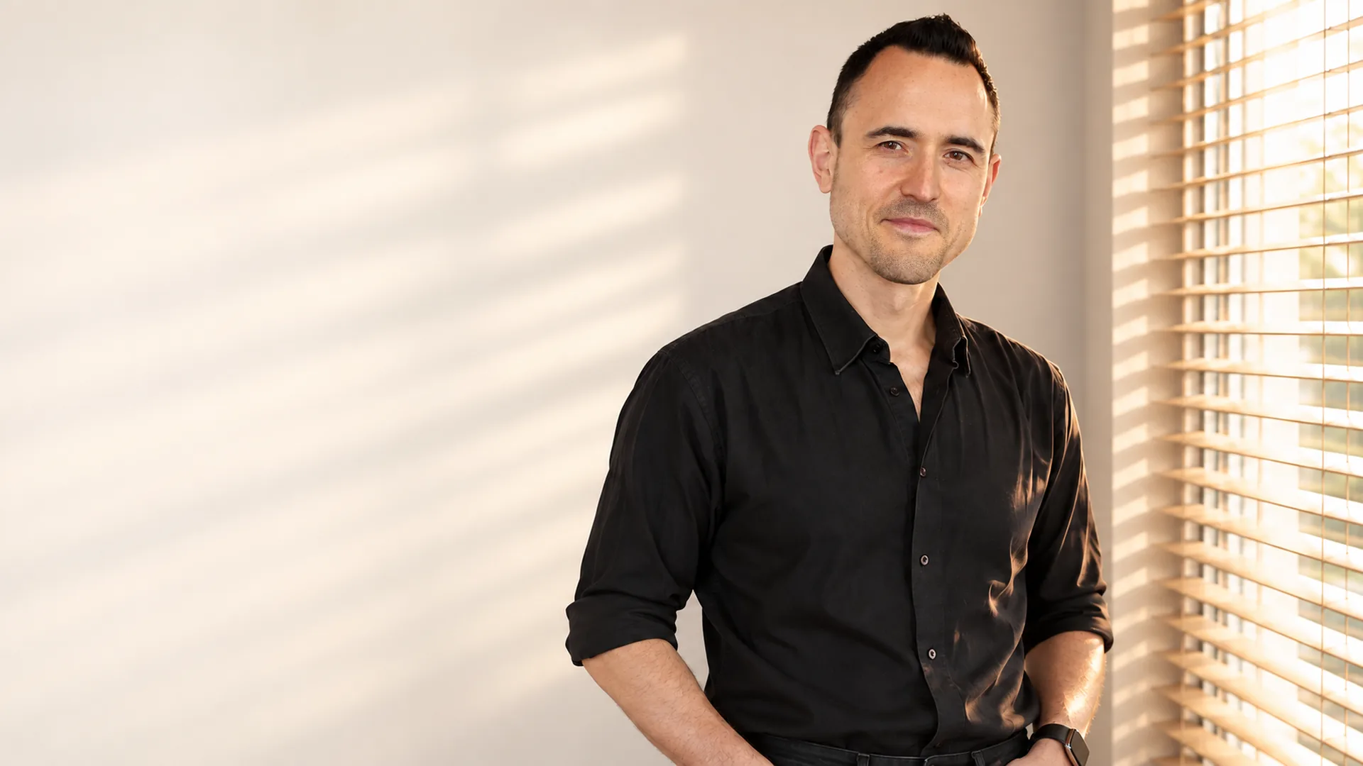 Julien Klein, chemise noire, sourire discret, debout près d'une fenêtre à stores, dans la lumière chaude de fin de journée.
